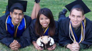 Three students wearing graduation regalia and laying on the grass