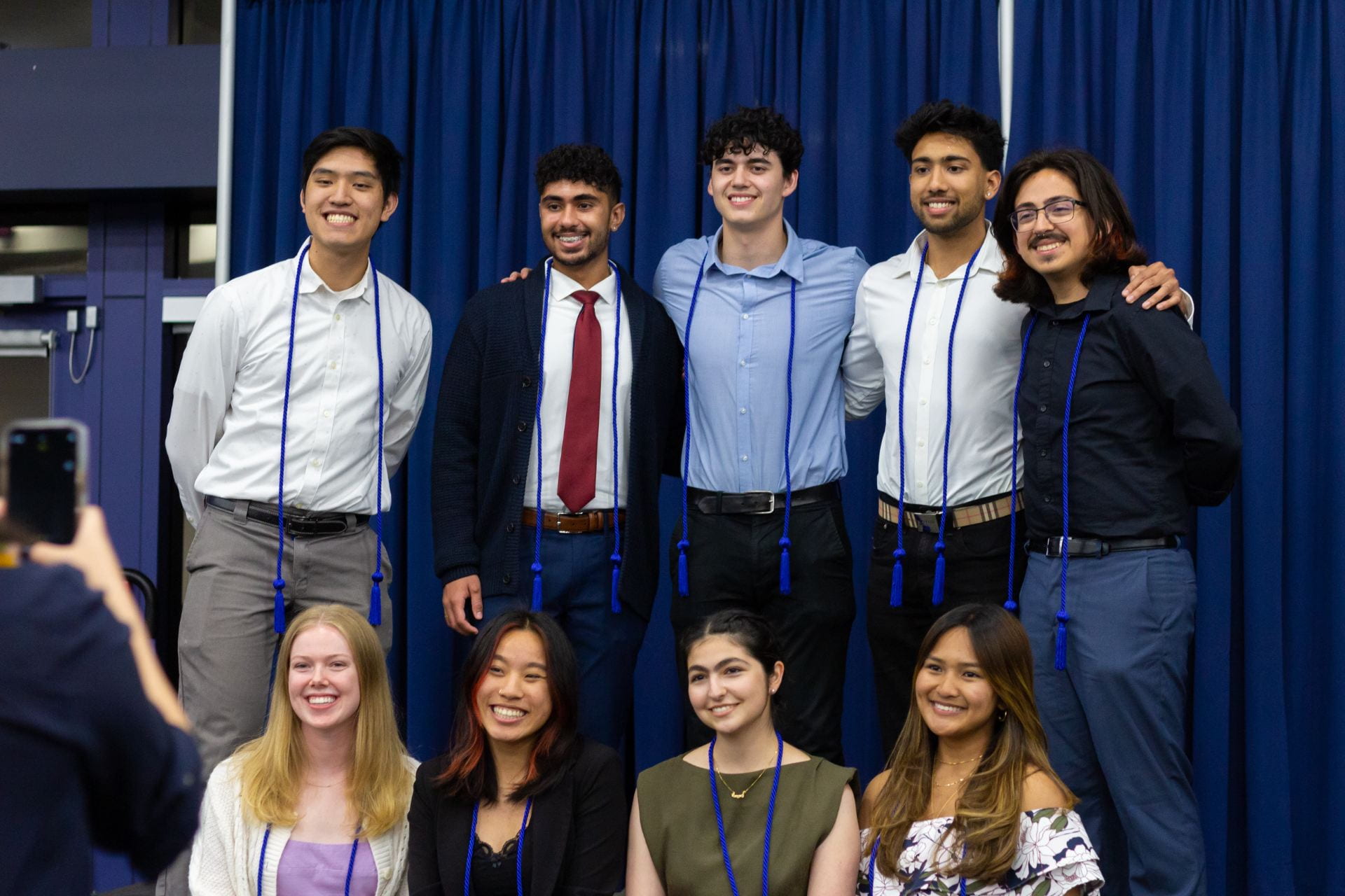 9 honors students pose with their blue honors coords and get into two rows to take a photo in front of a matching blue curtain
